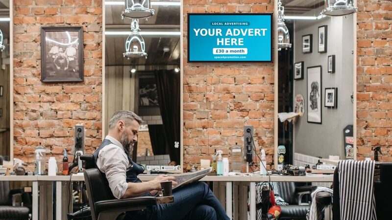 A Space4Promotion screen on the wall of a barbershop while a customer waits in the chair